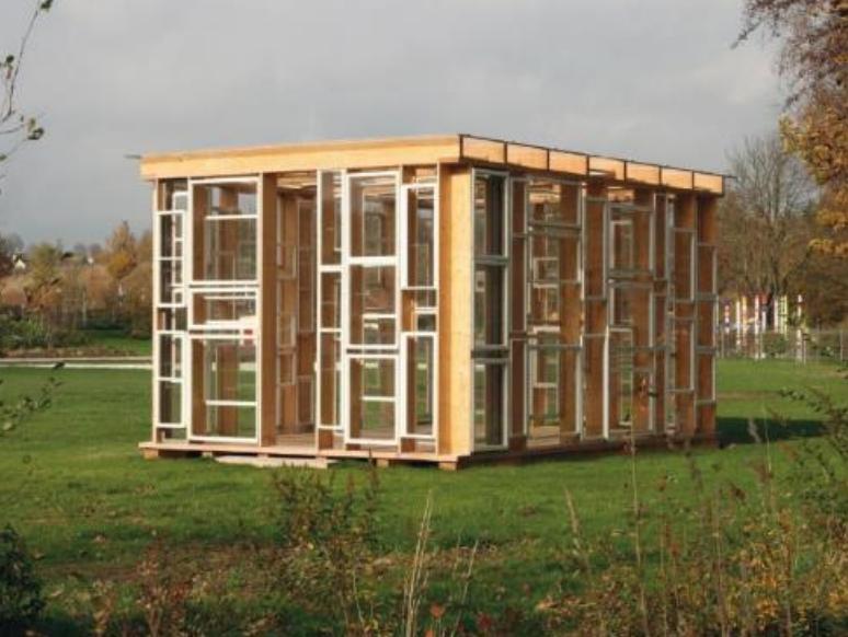 Kubusartiger Holzpavillon aus mehreren Fensterrahmen auf grüner Wiese